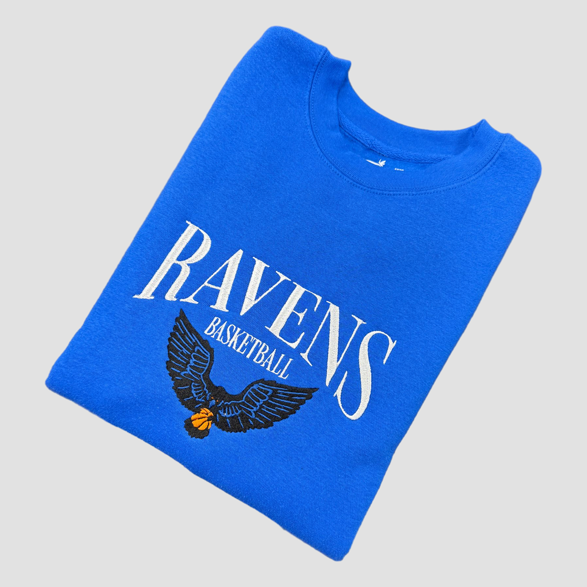 Folded blue sweatshirt with 'Ravens Basketball' text and eagle design on a gray background