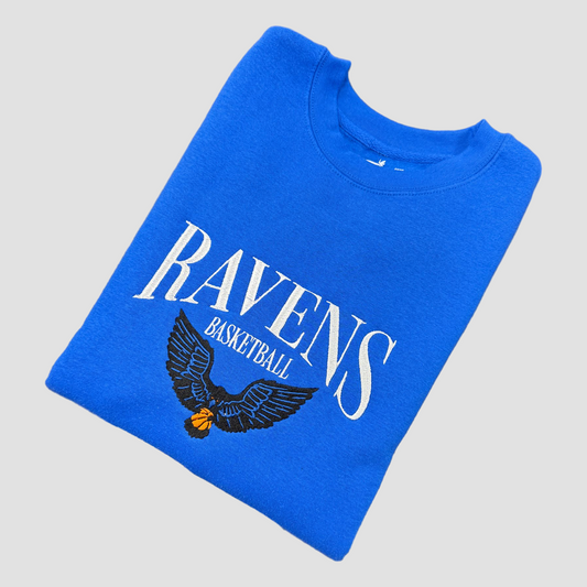 Folded blue sweatshirt with 'Ravens Basketball' text and eagle design on a gray background